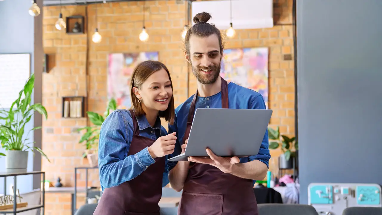 Young man and woman small business owners in aprons using a laptop inside their restaurant café.