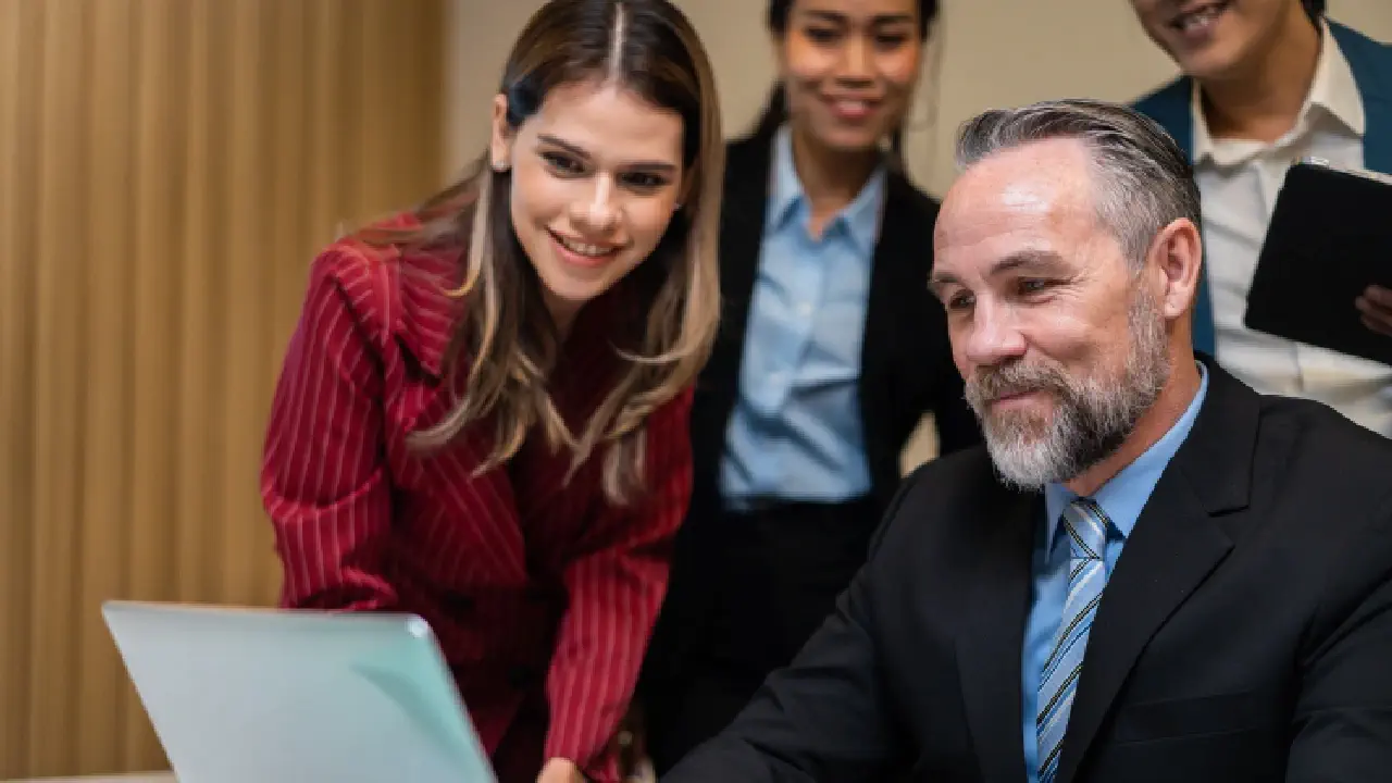 A man in a suit sits in front of a laptop, surrounded by his co-workers, representing a marketing team navigating the company's high-authority website