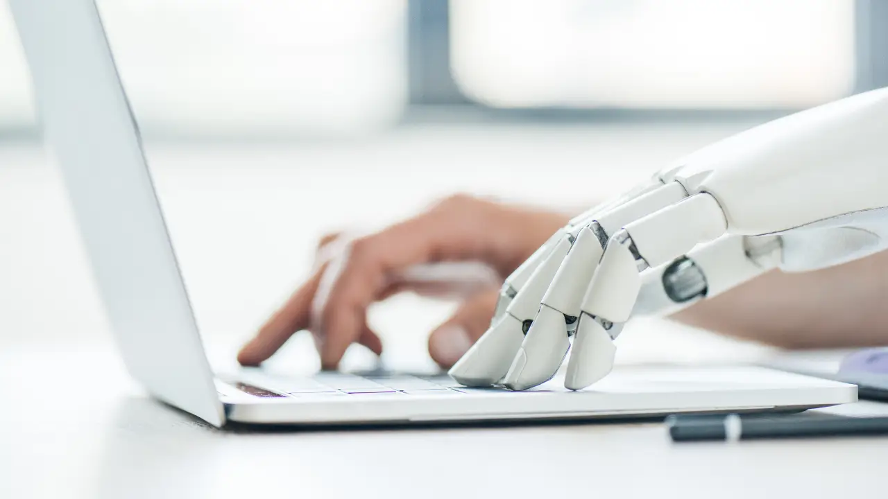 A close-up of a human hand and a robotic hand typing on a laptop keyboard together, symbolizing human-AI collaboration.